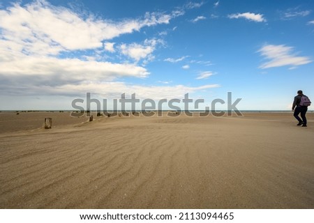 Backpacker walks along beach at Dunkirk with wooden poles of the sea defences and white clouds in a blue sky.