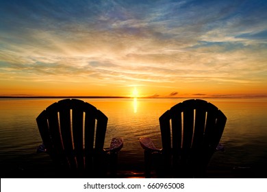  The Backs Of Two Adirondack Chairs Waiting At The End Of A Dock To Enjoy The Early Morning Summer Sunrise