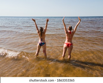 Backside aerial view: Two women in swimsuits stand and rise hands in sea water