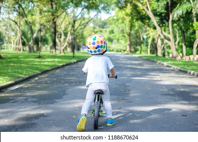 Backside Of Cute Asian 2 Years Toddler Boy Child Wearing Safety Helmet Learning To Ride First Balance Bike In Sunny Summer Day, Kid Cycling At Park, Explore & Appreciate Nature With Toddlers Concept