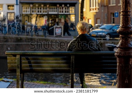 Backside of a old man on wooden bench with blurred view along the canal, The senior guy sitting and relaxing, Hobby and leisure activity of retirement age, Amsterdam capital city of the Netherlands.