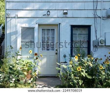 the backside of a small ran down white and blue house with peeling paint and old window with utilities and sunflowers growing around the walkway and a pale yellow door 