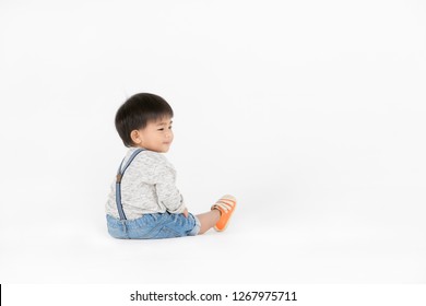 Backside of studio portrait of cute, adorable, Asian toddler boy wearing denim overalls, long sleeve T-shirt, orange shoes, turning his head to right, smiling, on isolated white background