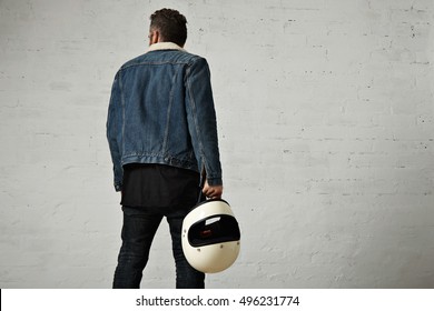 Backside view of brutal young motor biker wears shearling denim jacket and black blank henley shirt, walkinf away and holds vintage beige motorcycle helmet, isolated in center of white brick wall - Powered by Shutterstock