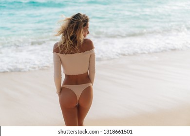 Backside view of girl with sexy booty in pink bikini resting on deserted beach. Beautiful model in swimwear walks along white sand on tropical island