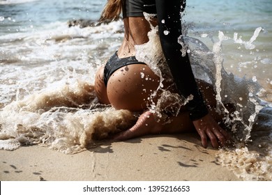 Backside view of girl with sexy booty in black bikini resting on deserted beach. Beautiful model in swimwear sitting on the sand on tropical island near big waves