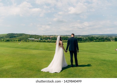 Backside view newlyweds couple holds hands each other and walks on green golf club glade. Bride in white wedding dress, groom in black business suit. Concept of just married - Powered by Shutterstock