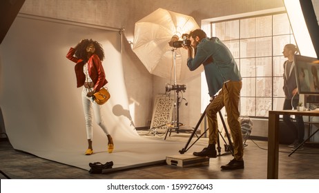 Backstage Of The Photo Shoot: Photographer Taking Photos Of Beautiful Black Model With Professional Camera. Fashion Magazine Studio Photoshoot. Golden Hour Shot.