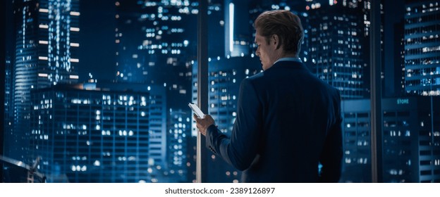 Backview of Handsome Caucasian Businessman in Stylish Suit Standing Next to Window in Big City Office with Skyscrapers Late At Night. Male CEO Using Smartphone To Check Work Emails. - Powered by Shutterstock