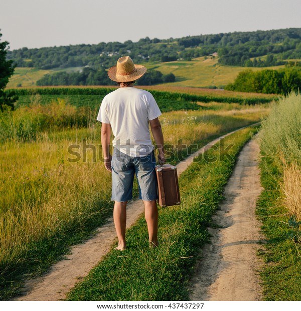 Backview of male caring old valise over blue sky\
outdoors background. Man wearing hat with suitcase walking away\
through wheat field.