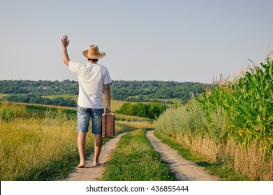 Backview Of Male Holding Old Valise Over Blue Sky Outdoors Background. Man Wearing Hat With Suitcase Walking Away Through Wheat Field.