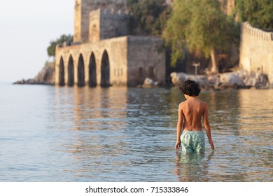 Backview Of Portrait Of Boy With Dark Hair Wearing Swimwear Standing In Water Near Alanya Medieval Shipyard In Mediterrarian Sea