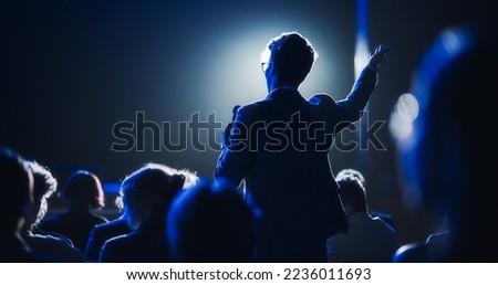 Backview of a Stylish Young Businessman in a Dark Crowded Auditorium at a Startup Summit. Young Man Talking to a Microphone During a Q and A session. Entrepreneur Happy with Event Speaker.