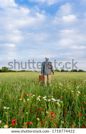 Backview of unrecognized male on blue sky day outdoors farmland nature background. Person holding old suitcase, relaxing hope freedom. Traveling away or coming back scene, blooming flowers