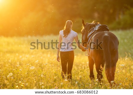 Backview of young woman walking with her horse in evening sunset light. Outdoor photography with fashion model girl. 