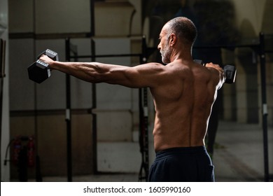 Backward View Of A Muscular Shirtless Mature Older Bodybuilding Athlete With Balding Gray Hair Does Side Lateral Dumbbell Raises In A Gym