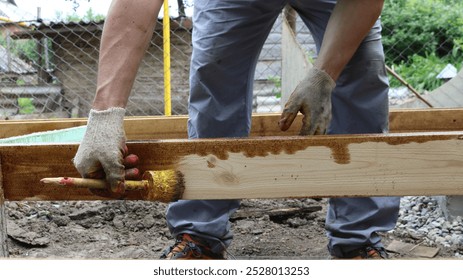 In the backyard a construction man in gray work pants and work gloves is painting a wooden beam. In his hand he holds a brush covered with varnish or brown paint.  - Powered by Shutterstock