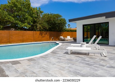 Backyard of elegant house in the Highland Lakes neighborhood of Miami, short grass, white walls, mirrored windows, swimming pool, sidewalk, wooden fence, black tiles, blue sky, palms, sun loungers - Powered by Shutterstock