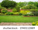 Backyard English cottage garden, colorful flowering plant and green grass lawn, brown pavement and orange brick wall, evergreen trees on background, in good care maintenance landscaping in park 
