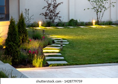 backyard of the mansion with a flowerbed and a lawn of green grass with a marble walkway of square tiles in the evening with a garden lighting with decorative ground lamps illuminating a warm light.