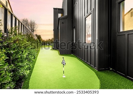 A backyard mini golf course with artificial turf next to a modern black house at sunset.