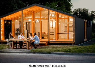 Backyard of the modern wooden house decorated with lights with friends dining at the table at the evening