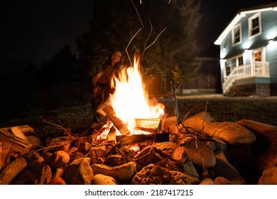  Backyard Of A Wooden House At Night. A Woman Covered With A Plaid Sits By The Fire. 