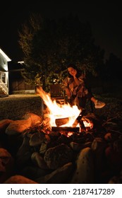  Backyard Of A Wooden House At Night. A Woman Covered With A Plaid Sits By The Fire. 