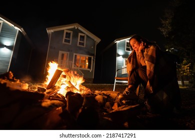 Backyard Of A Wooden House At Night. A Woman Covered With A Plaid Sits By The Fire. 