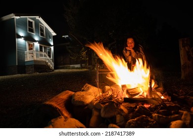  Backyard Of A Wooden House At Night. A Woman Covered With A Plaid Sits By The Fire. 