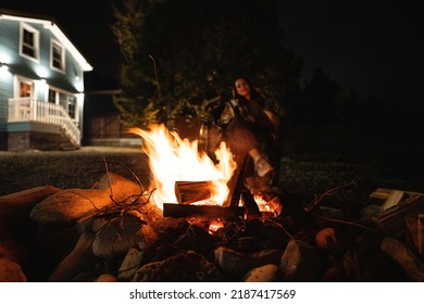  Backyard Of A Wooden House At Night. A Woman Covered With A Plaid Sits By The Fire. 