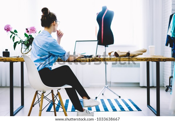Bacl view of charming female entrepreneur sitting at\
desktop with garment equipments and analyzing information from\
laptop computer with blank screen for your fashion advertising\
connected to wifi