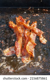 Bacon Frying In Grease On An Outdoor Camping Grill. The Pieces Are Piled Together In The Center.