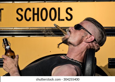 Bad Boys do not like school: Tough guy with sparrow beard, undercut and blue jeans leaning against his chopper bike in front of a yellow, American school bus. He is smoking cigar and drinking beer.