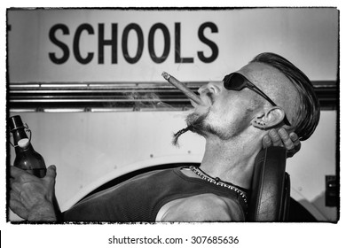 Bad Boys Do Not Like School: Tough Guy With Sparrow Beard, Undercut And Blue Jeans Sitting On His Chopper Bike In Front Of A Yellow, American School Bus. He Is Smoking Cigar And Drinking Beer.