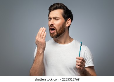 Bad breath. Handsome middle aged man checking his breath with his hand, blowing to it, standing over grey background. Bad smell from the mouth, toothache, having problems with teeth - Shutterstock ID 2255315769