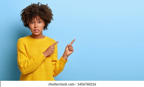 Bad choice. Dissatisfied dark skinned young lady points right nervously, has worried expression, purses lips, has curly hairstyle, wears yellow sweater, shows offer, isolated on blue background.