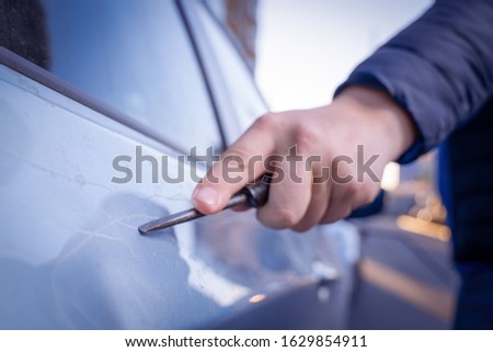 Bad guy scratching the car door with a screwdriver in the parking lot on the street. Damage of property from revenge for treason or betrayal, or threat. Auto insurance fraud or vandalism. 