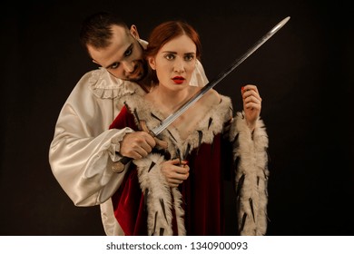 Bad Guy In A White Shirt Wants To Kill A Young Beautiful Girl In A Red Dress On A Black Background. Emotional Frightened Face. Scene Of The Medieval Ago.