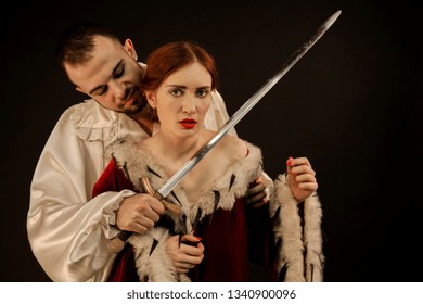 Bad Guy In A White Shirt Wants To Kill A Young Beautiful Girl In A Red Dress On A Black Background. Emotional Frightened Face. Scene Of The Medieval Ago.