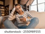 Bad News Concept. Upset Confused Woman Holding Smartphone, Looking At Mobile Phone Screen With Worried Expression, Touching Head, Sad Adult Female Reading Unplesant Message Sitting On Couch