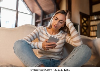 Bad News Concept. Upset Confused Woman Holding Smartphone, Looking At Mobile Phone Screen With Worried Expression, Touching Head, Sad Adult Female Reading Unplesant Message Sitting On Couch