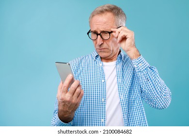 Bad News Concept. Upset Mature Man Holding Smartphone, Looking At Mobile Phone Screen With Worried Expression, Touching Glasses, Sad Adult Male Reading Unplesant Message, Blue Studio Background