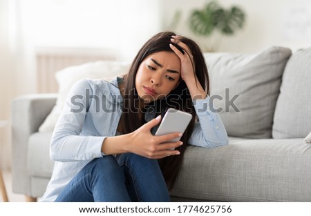 Bad News. Depressed Asian Girl Looking At Smartphone Screen At Home With Sad Face Expression, Sitting On Floor Near Couch, Closeup