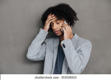 Bad news. Frustrated young African man talking on mobile phone and holding hand in hair while standing against grey background