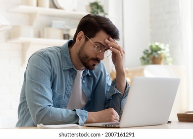 Bad News, Lack Of Understanding Of New App, Info Overload, Device Malware Concept. Young Man In Glasses Sit At Table Looks At Laptop Screen Touch Forehead Feels Confused Has Business Or Pc Problems