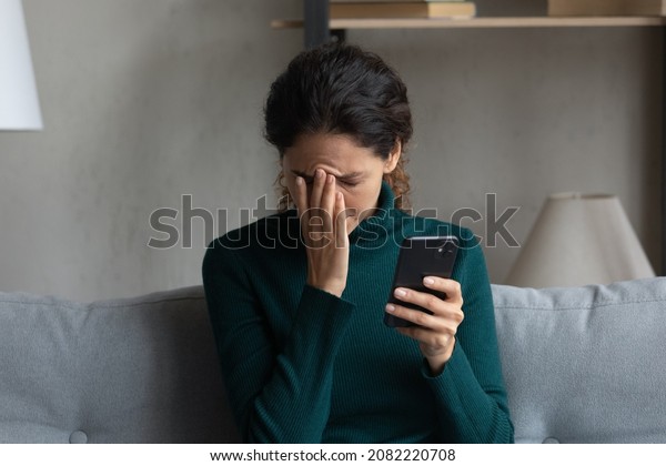 Bad news on screen. Confused frustrated young\
latin lady cover face with palm turn away from cell seeing\
important call missed. Desperate millennial woman get message on\
phone about dismissal from\
job