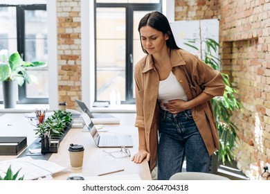 Bad period. Full length view of the caucasian woman suffering with menstrual stomach ache or feeling hurting stomach in her office  - Shutterstock ID 2064704318