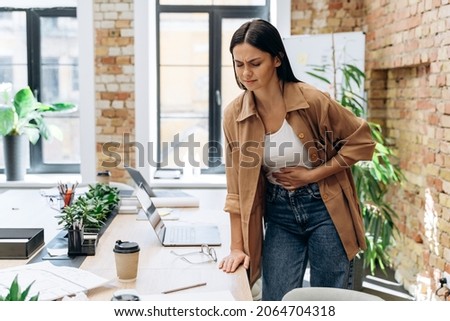 Bad period. Full length view of the caucasian woman suffering with menstrual stomach ache or feeling hurting stomach in her office 