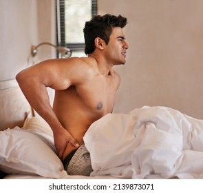 Bad Quality Sleep Leaves Him Feeling Stiff And Sore. Shot Of A Young Man Waking Up In Bed With Back Pain.